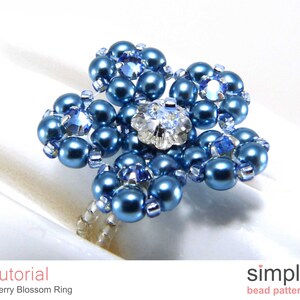 Könnte beinhalten: Ein blauer Perlenring mit Blumenmuster. Der Ring ist aus kleinen blauen Perlen und einem größeren Kristall in der Mitte gefertigt. Der Ring liegt auf einer weißen Oberfläche.