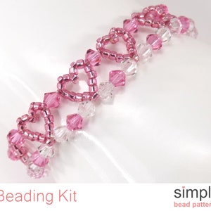 Op de afbeelding: Een roze en transparante kralenarmband met een hartvormig ontwerp. De armband is gemaakt van kleine kraaltjes en grotere kristallen kralen. De armband ligt op een witte achtergrond.