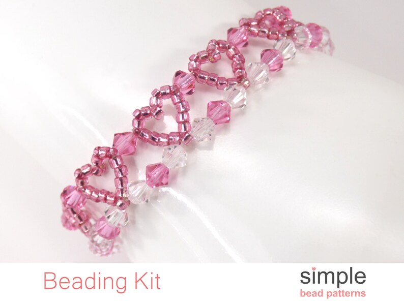 Op de afbeelding: Een roze en transparante kralenarmband met een hartvormig ontwerp. De armband is gemaakt van kleine kraaltjes en grotere kristallen kralen. De armband ligt op een witte achtergrond.