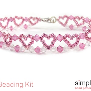 Op de afbeelding: Een armband van roze en heldere kristallen kralen met een hartvormig ontwerp. De armband is gemaakt van rocailles en heeft een zilveren sluiting. Kralenset.