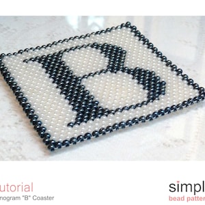 Puede incluir: Un posavasos de cuentas blancas y negras con la letra "B" en el centro. El posavasos está hecho con cuentas pequeñas y redondas.
