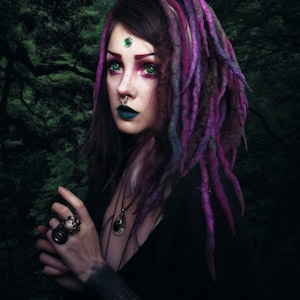 May include: A woman with purple and pink dreadlocks, green eyes, and a green gemstone on her forehead. She is wearing a black top and has a silver nose ring and a silver necklace with a dark gemstone pendant.