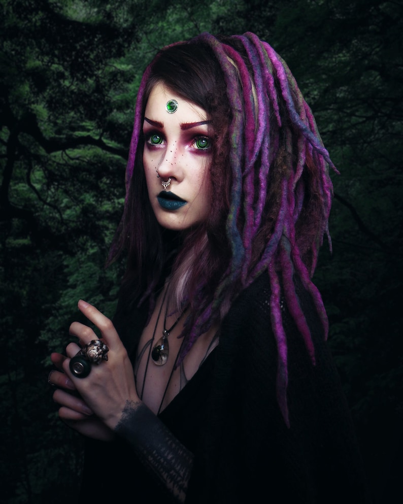 May include: A woman with purple and pink dreadlocks, green eyes, and a green gemstone on her forehead. She is wearing a black top and has a silver nose ring and a silver necklace with a dark gemstone pendant.