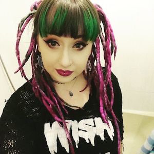 May include: A person with green and pink dreadlocks, wearing a black shirt with the word "WISH" printed on it in white. The person has a nose ring and a lip piercing.