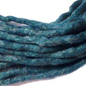 Könnte beinhalten: Nahaufnahme eines Bündels teal-farbener Dreadlocks. Das Kunsthaar hat ein strukturiertes Aussehen mit subtilen irisierenden Strähnen, die eingewebt sind und einen schimmernden Effekt erzeugen. Die Dreadlocks sind eng gedreht und scheinen handgefertigt zu sein.