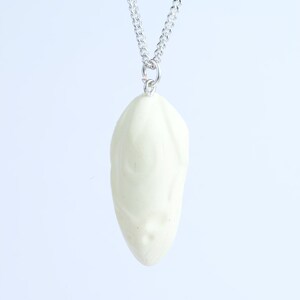 May include: A cream-colored pendant necklace featuring a sculpted face design. The pendant is suspended from a silver-colored chain. The face has subtle details, and the overall aesthetic is minimalist and artistic.