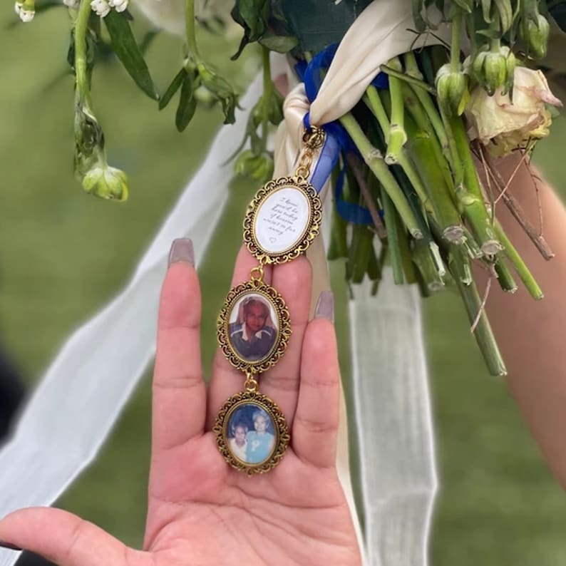 May include: A gold-toned metal memorial charm featuring three oval frames attached to a bouquet with white and blue ribbons. The top frame contains text, while the two lower frames hold photographs of people. The frames have an ornate filigree border.