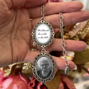 May include: A silver chain necklace with two oval-shaped charms. The top charm has a white background with the text "She believed she could... so she did!" The bottom charm has a black and white photo of an older woman.