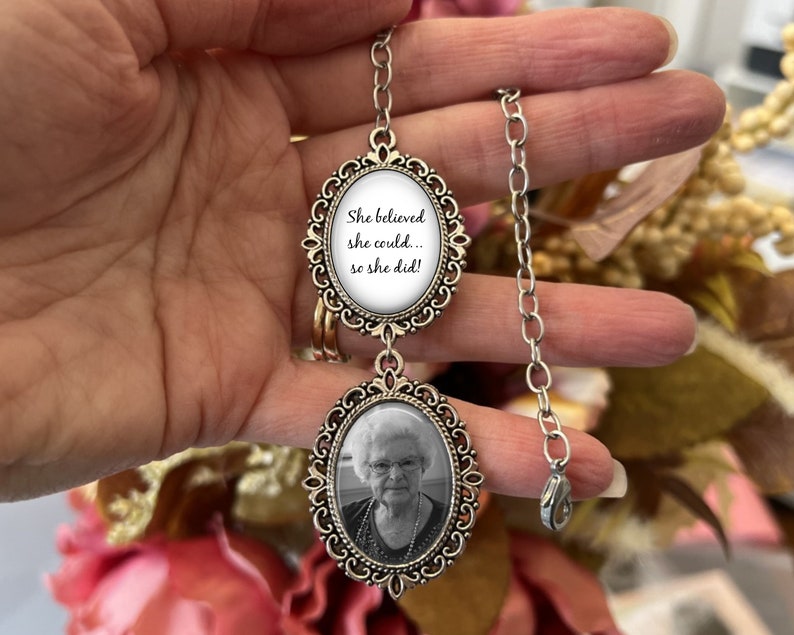 May include: A silver chain necklace with two oval-shaped charms. The top charm has a white background with the text "She believed she could... so she did!" The bottom charm has a black and white photo of an older woman.