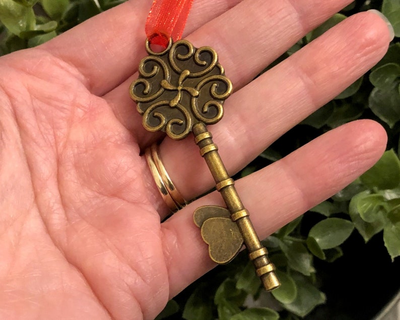 May include: An antique-style key pendant with an ornate, swirling design on the head and a butterfly-shaped detail. The key is a bronze colour and is attached to a red ribbon. The key is held in a hand.