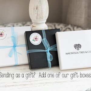 May include: Three gift boxes with blue ribbon and a magnolia flower logo. The boxes are white, black, and white with the text "MAGNOLIA TREE & CO."