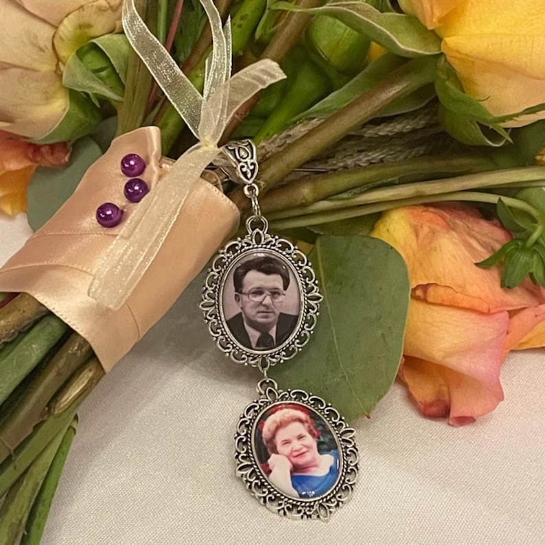 May include: A silver-toned metal pendant featuring two oval frames with ornate filigree borders. The top frame contains a black and white portrait of a man wearing glasses, while the bottom frame holds a color portrait of a woman.