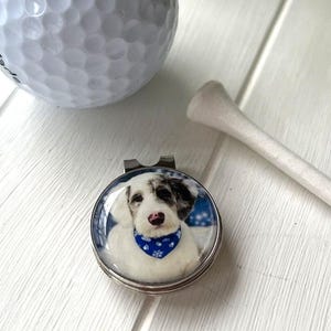 May include: A golf ball, tee, and a silver-colored magnetic golf ball marker with a clear, domed top. The marker features a close-up image of a white and black dog wearing a blue bandana. The golf ball is white.