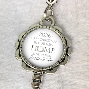 May include: A silver key-shaped ornament with a circular central design. The design includes the text "2026 - FIRST CHRISTMAS IN OUR NEW HOME" and the address "21 Santa Way Jessie & Tom". A floral wreath design is also visible.