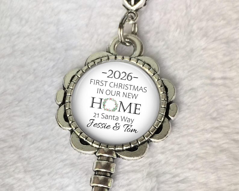 May include: A silver key-shaped ornament with a circular central design. The design includes the text "2026 - FIRST CHRISTMAS IN OUR NEW HOME" and the address "21 Santa Way Jessie & Tom". A floral wreath design is also visible.