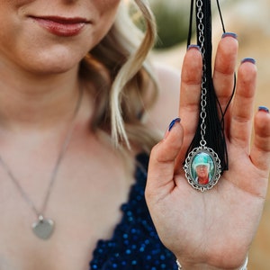 May include: A woman in a blue sequined dress holds a graduation tassel with a silver charm featuring a photo of a person wearing a green hat.