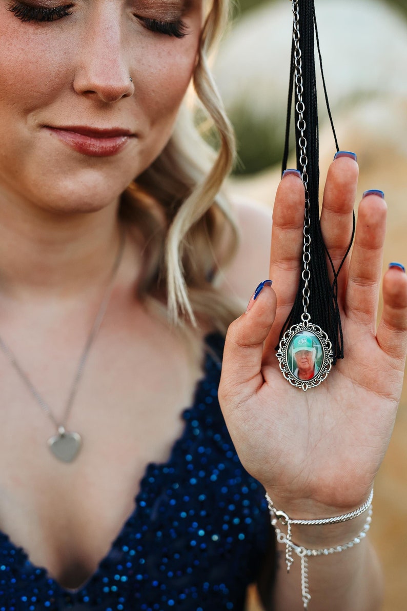 May include: A woman in a blue sequined dress holds a graduation tassel with a silver charm featuring a photo of a person wearing a green hat.