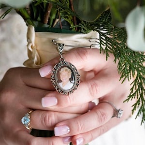 May include: A silver-toned oval photo charm pendant featuring a portrait of a person with blonde hair. The pendant is attached to a silver safety pin, which is secured to the cream-colored ribbon wrapping the stems of a green floral bouquet.