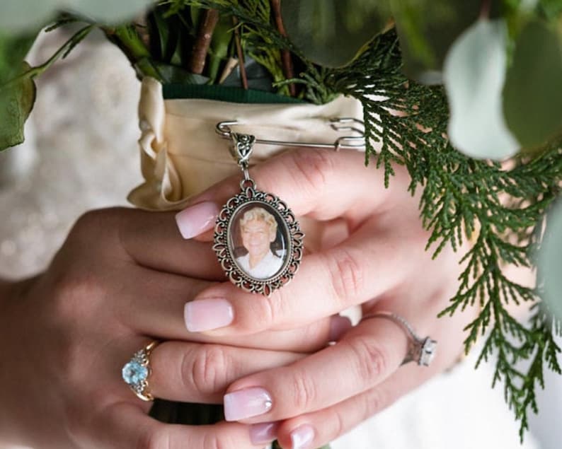 May include: A silver-toned oval photo charm pendant featuring a portrait of a person with blonde hair. The pendant is attached to a silver safety pin, which is secured to the cream-colored ribbon wrapping the stems of a green floral bouquet.