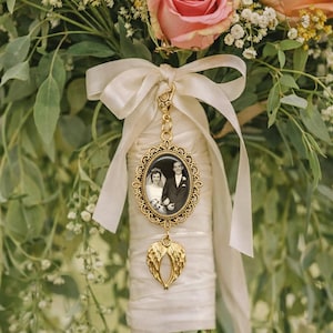 May include: A gold-toned oval photo charm featuring a black and white wedding portrait of a couple, suspended from a cream-coloured satin ribbon bow. Below the oval frame hangs a small gold-toned charm shaped like a pair of angel wings.