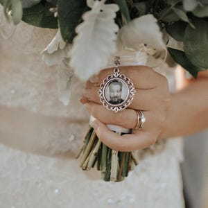 May include: A white bridal bouquet with a silver locket charm featuring a black and white photo of a man. The locket is attached to a white ribbon.