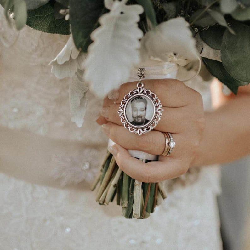 May include: A white bridal bouquet with a silver locket charm featuring a black and white photo of a man. The locket is attached to a white ribbon.