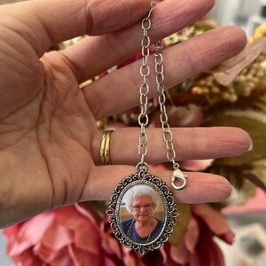 May include: Silver chain with a silver oval locket featuring a photo of an older woman with white hair. The locket has a decorative border.