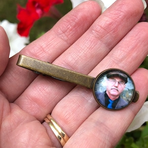 May include: A bronze tie clip with a round glass insert featuring a photo of a man wearing glasses and a hat.