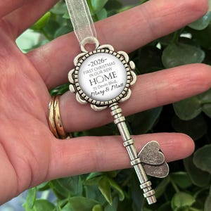May include: A silver-coloured key-shaped Christmas ornament with a floral design around a circular insert. The insert reads "-2026 FIRST CHRISTMAS IN OUR NEW HOME 72 Devin Blvd. Mary & Matt." A sheer ribbon is attached for hanging.