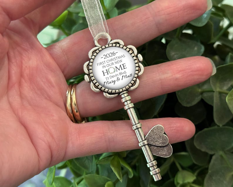 May include: A silver-coloured key-shaped Christmas ornament with a floral design around a circular insert. The insert reads "-2026 FIRST CHRISTMAS IN OUR NEW HOME 72 Devin Blvd. Mary & Matt." A sheer ribbon is attached for hanging.