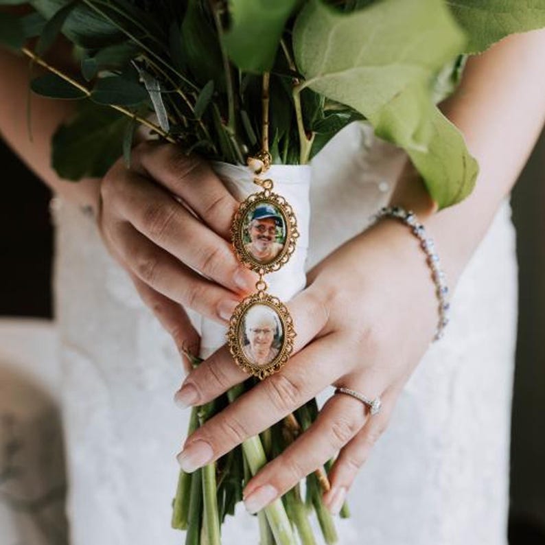May include: A gold-toned metal locket charm featuring two oval frames connected vertically. Each frame contains a small portrait photograph of an individual. The charm is attached to a white ribbon wrapped around a bouquet of green leaves and stems.