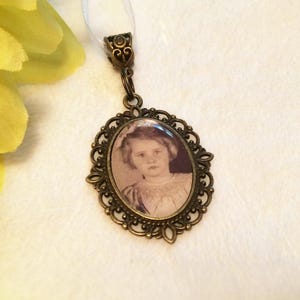 May include: An oval pendant featuring a sepia-toned photograph of a young child with short hair, encased in an ornate, antique bronze-colored metal frame with intricate filigree detailing and a matching decorative bail attached to a white ribbon.