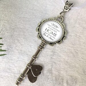May include: A silver-coloured key-shaped ornament with a round, floral-edged pendant. The pendant has the text "-2026- FIRST CHRISTMAS IN OUR NEW HOME 4 Blue Heron Way Bri & Steve" in black. A white ribbon is attached.