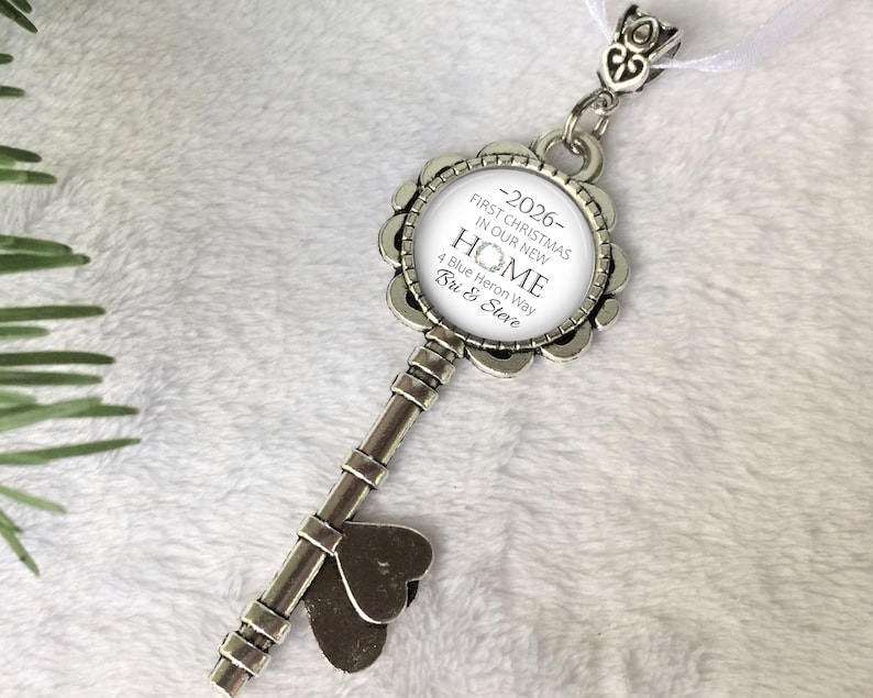 May include: A silver-coloured key-shaped ornament with a round, floral-edged pendant. The pendant has the text "-2026- FIRST CHRISTMAS IN OUR NEW HOME 4 Blue Heron Way Bri & Steve" in black. A white ribbon is attached.