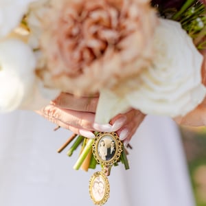 May include: A gold-toned metal photo charm featuring two oval frames with vintage black and white portraits, attached to a bouquet of white and tan roses. The top frame displays a couple, while the smaller dangling frame shows two individuals.