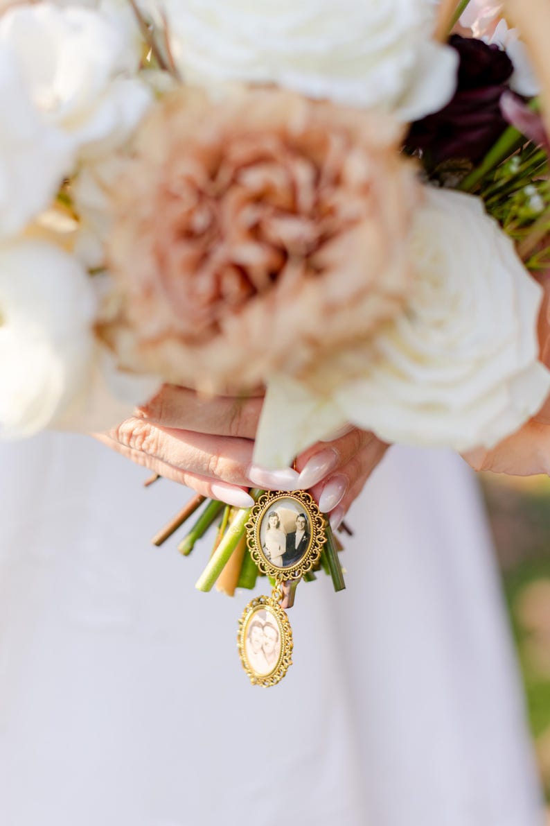 May include: A gold-toned metal photo charm featuring two oval frames with vintage black and white portraits, attached to a bouquet of white and tan roses. The top frame displays a couple, while the smaller dangling frame shows two individuals.