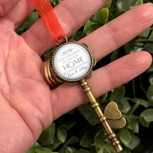 May include: A bronze-coloured key-shaped Christmas ornament with a red ribbon. The round pendant displays the text "-2026- FIRST CHRISTMAS IN OUR NEW HOME 405 Waterdown Way Kim & Alex". The key has a butterfly design.