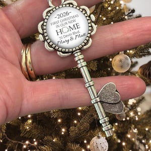 May include: Silver-coloured key-shaped Christmas ornament. The ornament has a circular centre with the text "-2026- FIRST CHRISTMAS IN OUR NEW HOME 72 Devin Blvd. Mary & Matt." The key has a butterfly design and is hanging from a green ribbon.