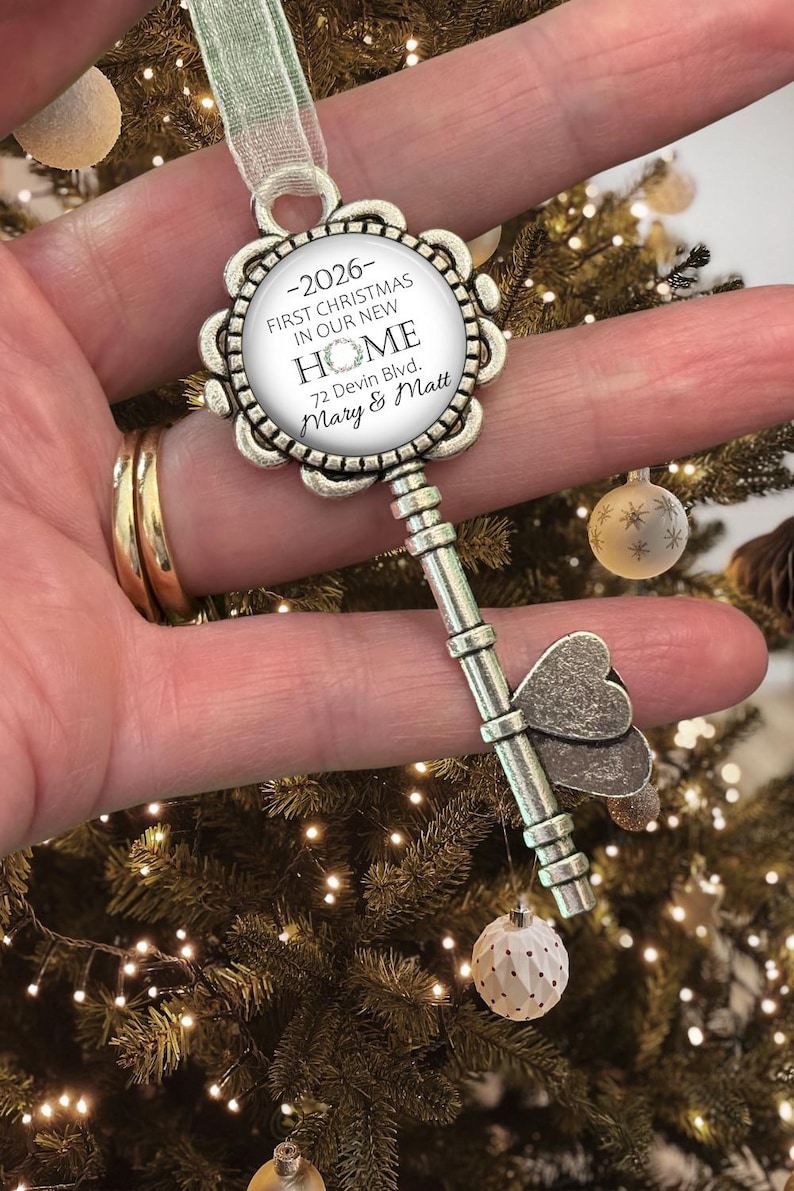 May include: Silver-coloured key-shaped Christmas ornament. The ornament has a circular centre with the text "-2026- FIRST CHRISTMAS IN OUR NEW HOME 72 Devin Blvd. Mary & Matt." The key has a butterfly design and is hanging from a green ribbon.
