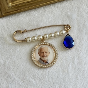 May include: A gold-tone safety pin brooch featuring a row of white faux pearls, a circular pendant containing a photograph of an older man with a white beard, and a single faceted teardrop-shaped blue crystal charm hanging from the side.