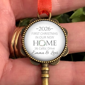 May include: A decorative key-shaped Christmas ornament with a circular pendant. The pendant has the text "2026 - First Christmas in our New Home", an address, and names. It is suspended by a red ribbon.