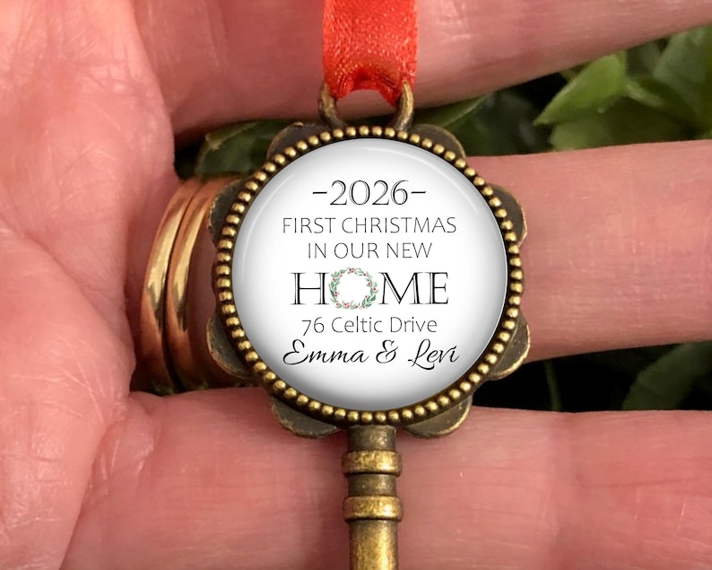 May include: A decorative key-shaped Christmas ornament with a circular pendant. The pendant has the text "2026 - First Christmas in our New Home", an address, and names. It is suspended by a red ribbon.
