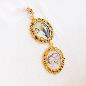 May include: A gold-toned metal pendant featuring two oval frames with intricate filigree borders. Each frame contains a black and white photograph of a couple. The pendant is attached to a white ribbon at the top, creating a hanging ornament.