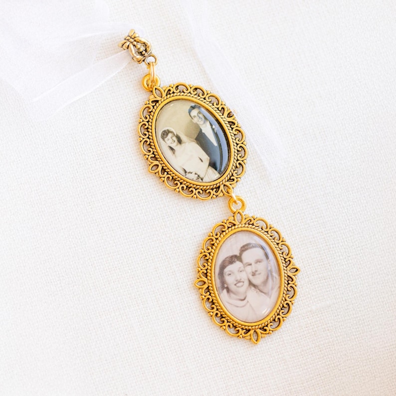 May include: A gold-toned metal pendant featuring two oval frames with intricate filigree borders. Each frame contains a black and white photograph of a couple. The pendant is attached to a white ribbon at the top, creating a hanging ornament.