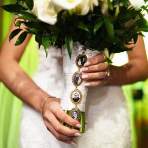 May include: A bridal bouquet featuring white calla lilies and green foliage, with a white ribbon-wrapped stem. Attached to the stem is a vertical gold-toned metal charm chain featuring four oval frames, each containing a small black and white photograph.