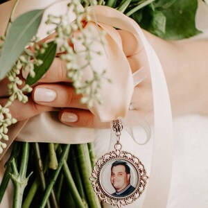 May include: A white bridal bouquet with a silver charm attached. The charm features a photo of a man in a black shirt.