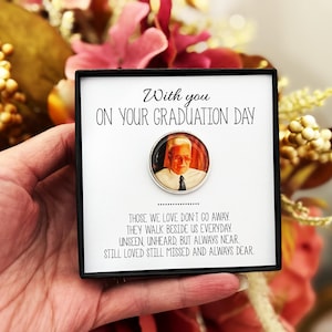May include: A black box with a white card that reads "With you ON YOUR GRADUATION DAY." A circular pin with a photo of a man is on the card. The text below reads "THOSE WE LOVE DON'T GO AWAY..."