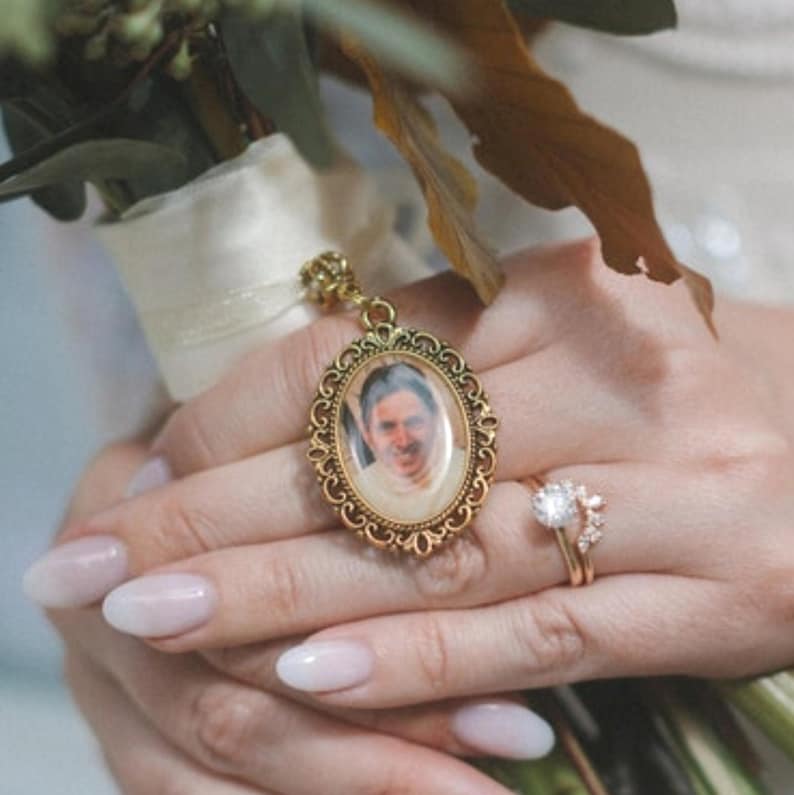 May include: An oval gold-toned metal locket featuring an intricate filigree border and a portrait of a person inside. The locket is attached to a cream-colored ribbon and is positioned alongside a gold ring with a clear round stone and side accents.