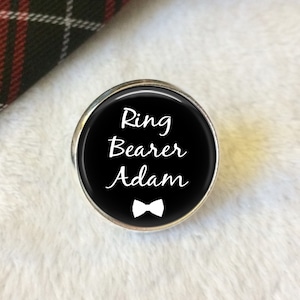 May include: A black and silver ring bearer button with white text that reads "Ring Bearer Adam" and a white bow tie.
