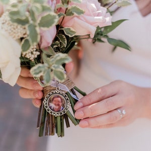 May include: A white bridal bouquet with pink roses and greenery, featuring a silver locket charm with a photo of a person inside.